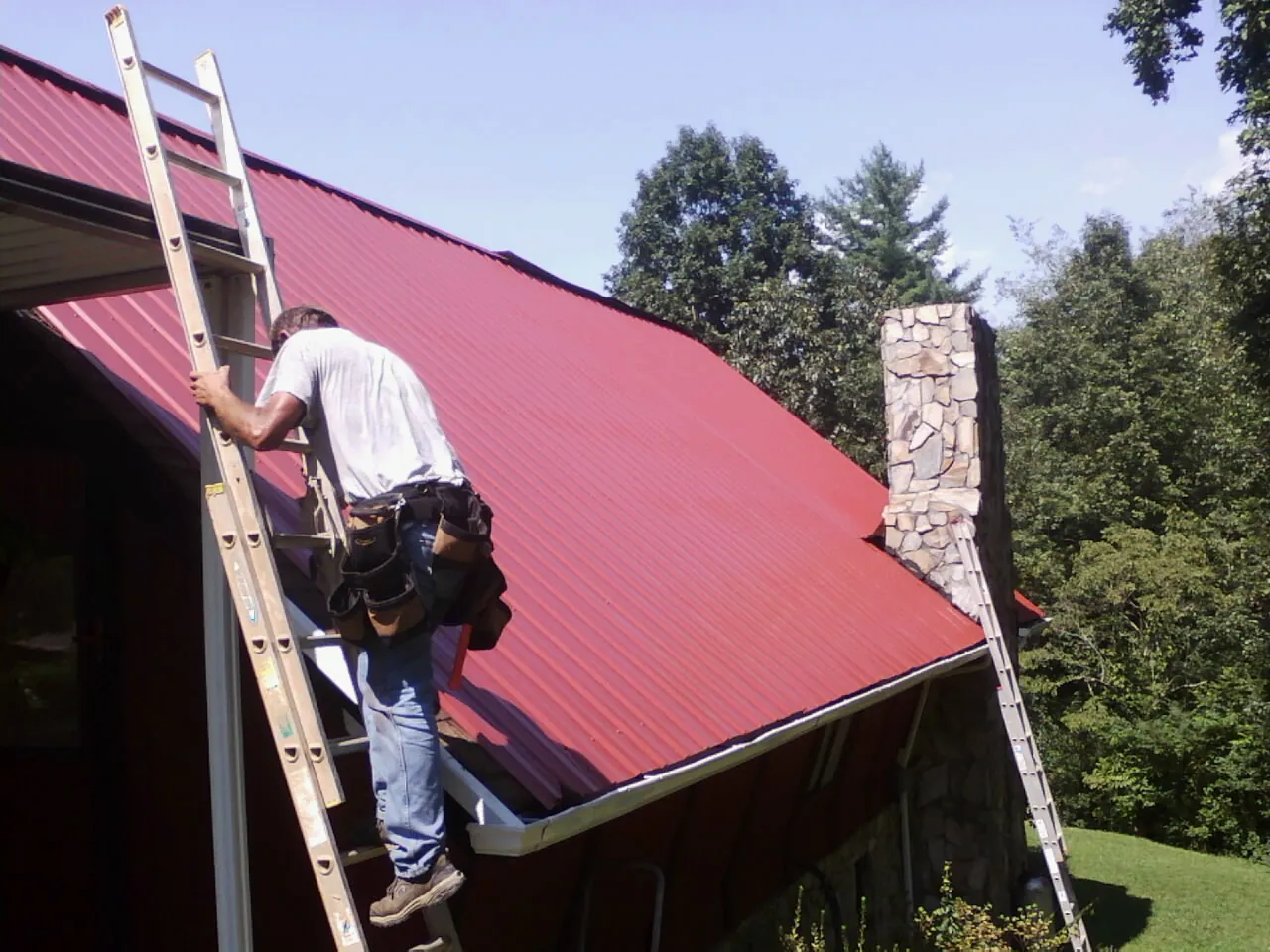 Roofer on a ladder beside a red metal roof and stone chimney — Soffit & Fascia Repair by Roofing Brothers in Weslaco